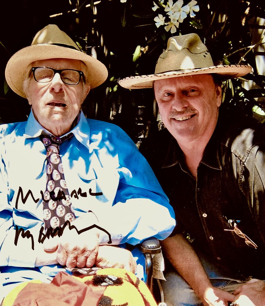 Ray Bradbury in a straw hat and bright shirt sitting beside Michael O'Kelly, also in a hat.