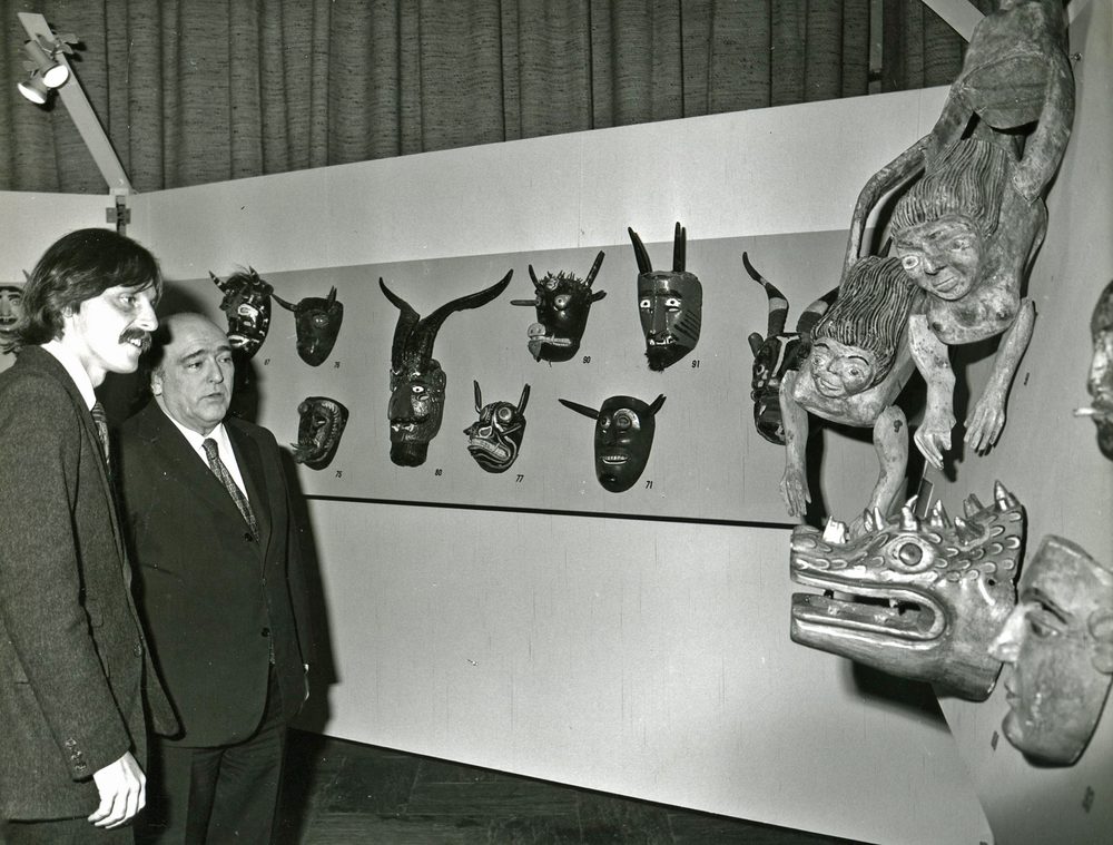 Black-and-white photograph of two men in suits looking at a wall of Mexican masks at an exhibition opening.
