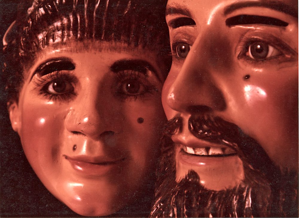 Two carved and painted Mexican dance masks, a pink-cheeked man with a black beard and a younger face beside him.
