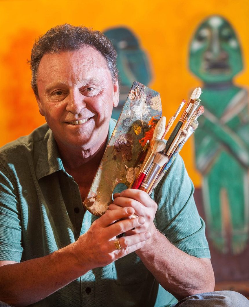 Michael O'Kelly in his studio, holding a paint-loaded palette and a fist of brushes, in front of an orange wall hung with two carved and painted figures from his collection.