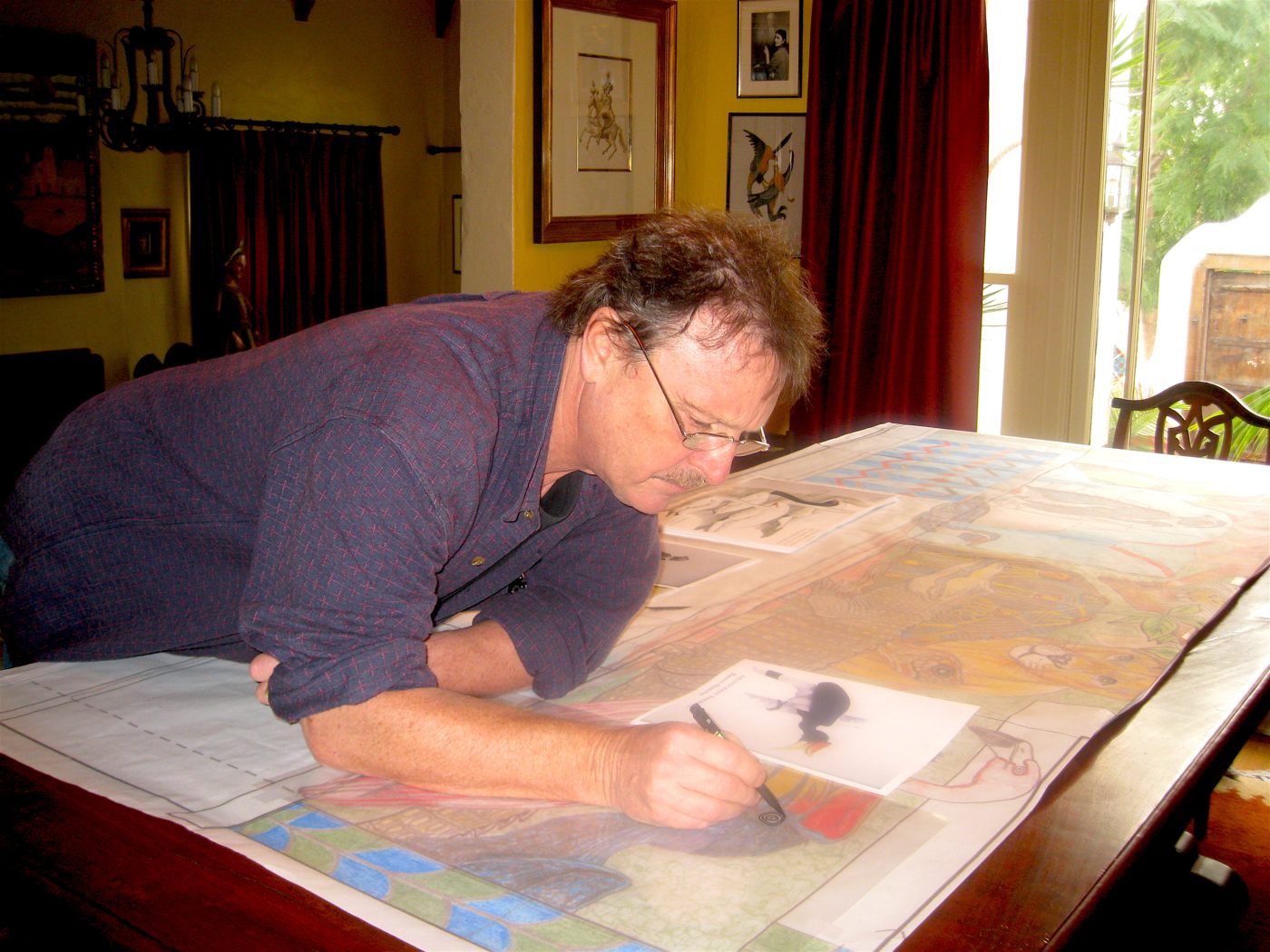 Michael O'Kelly leaning over a long table, drawing the full-size cartoon of the fountain wall with animal reference photographs laid beside him.