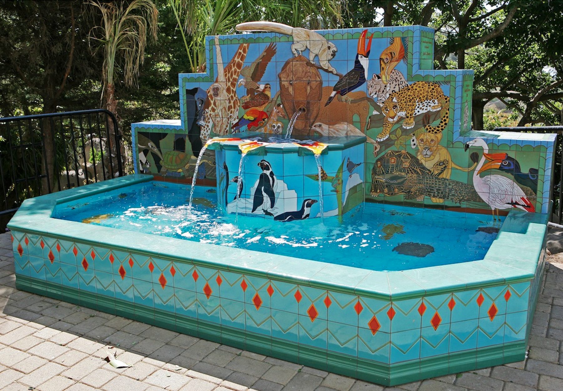 The Animal Kingdom Fountain at Santa Barbara Zoo: a stepped tiled wall crowded with animals &mdash; a giraffe, an elephant whose trunk spouts water, a toucan, a jaguar and a snow leopard, a flamingo and a crocodile &mdash; above a tiled block of penguins and a long turquoise pool bordered with red diamonds.
