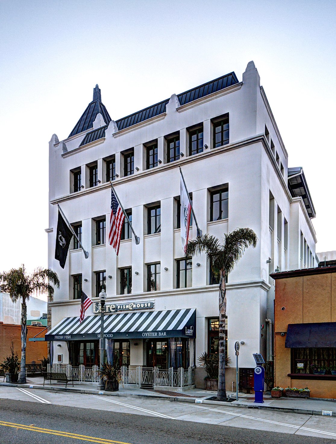 60 California Street, Ventura: a four-storey white building with a pointed corner tower and dark roof, flags over the street, and the striped awning of Lure Fish House at street level.