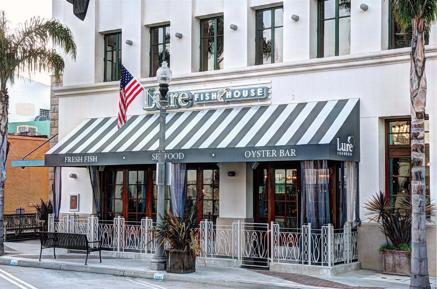 Street level at 60 California Street: Lure Fish House under a grey-and-white striped awning reading Fresh Fish, Seafood, Oyster Bar, behind a white iron railing, with a period street lamp and palm trees.