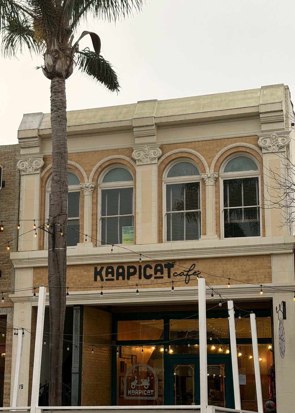 The Kaapicat Café building today: a cream and ochre facade with four arched upper windows above the Kaapicat sign and teal doors.