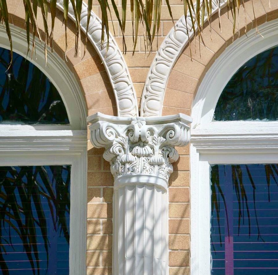 Close-up of a white Corinthian capital with a carved face, between two arched windows on the facade.