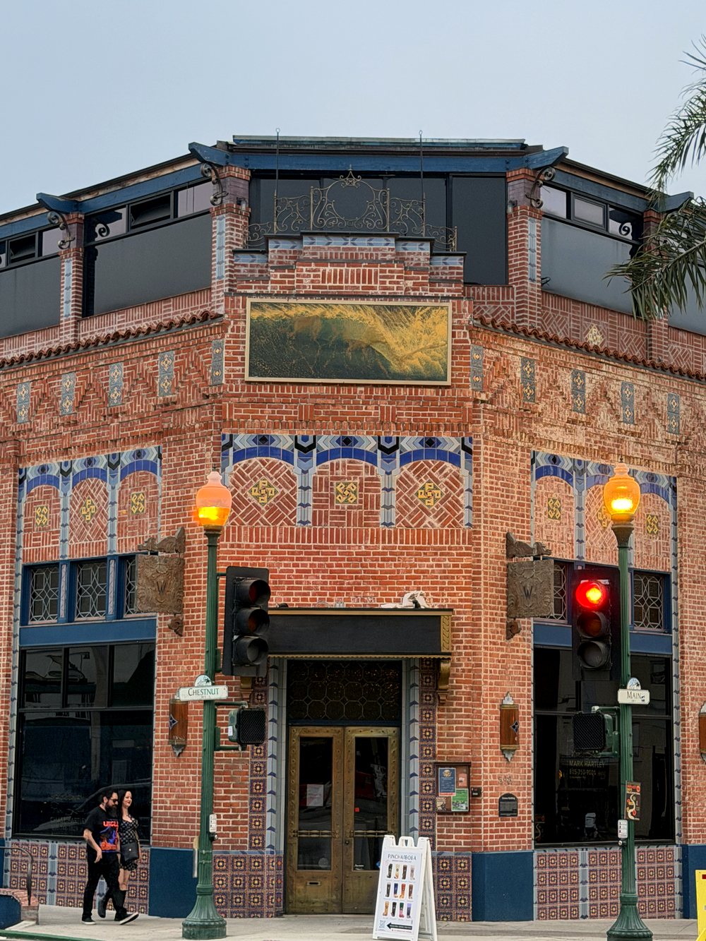 The Watermark corner entrance: brass doors under a gilded panel, framed by bands of blue tile arches and patterned brick, with period street lamps either side.