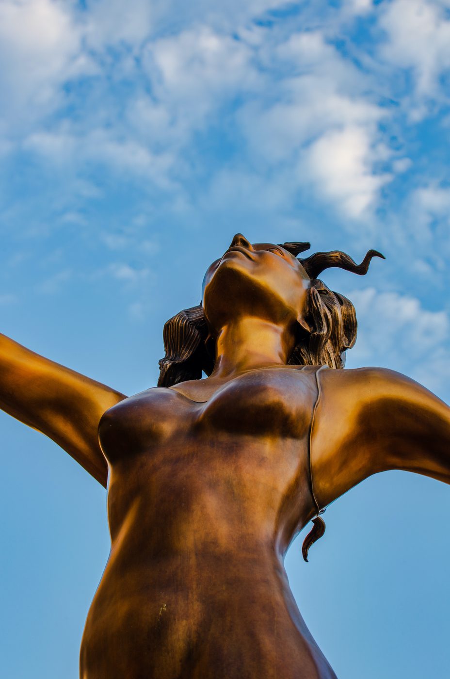 Looking up at the bronze dancer from below, arms spread wide against a sky of white clouds.