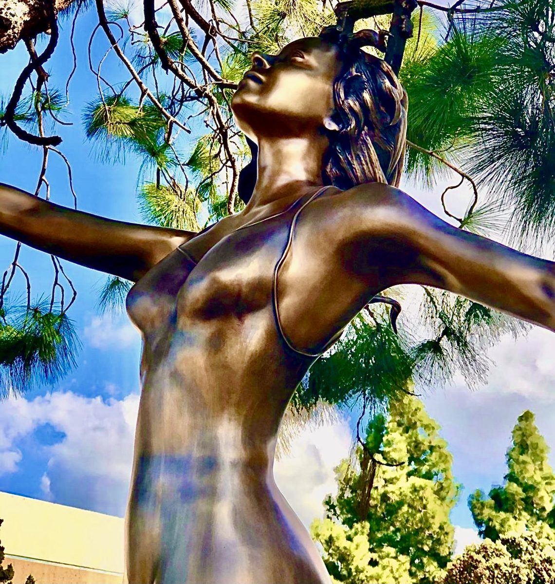 Close-up of the dancer's upturned face and outstretched arm, with pine trees and blue sky behind.