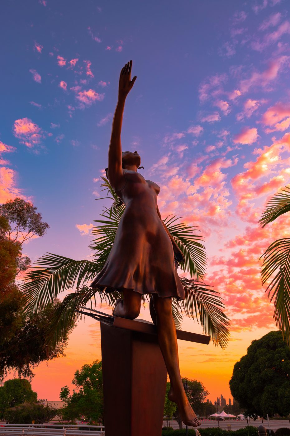 Stepping Out: a twelve-foot bronze figure of a dancer with one arm raised to the sky, silhouetted against a pink and violet sunset and palm trees at Ventura College.